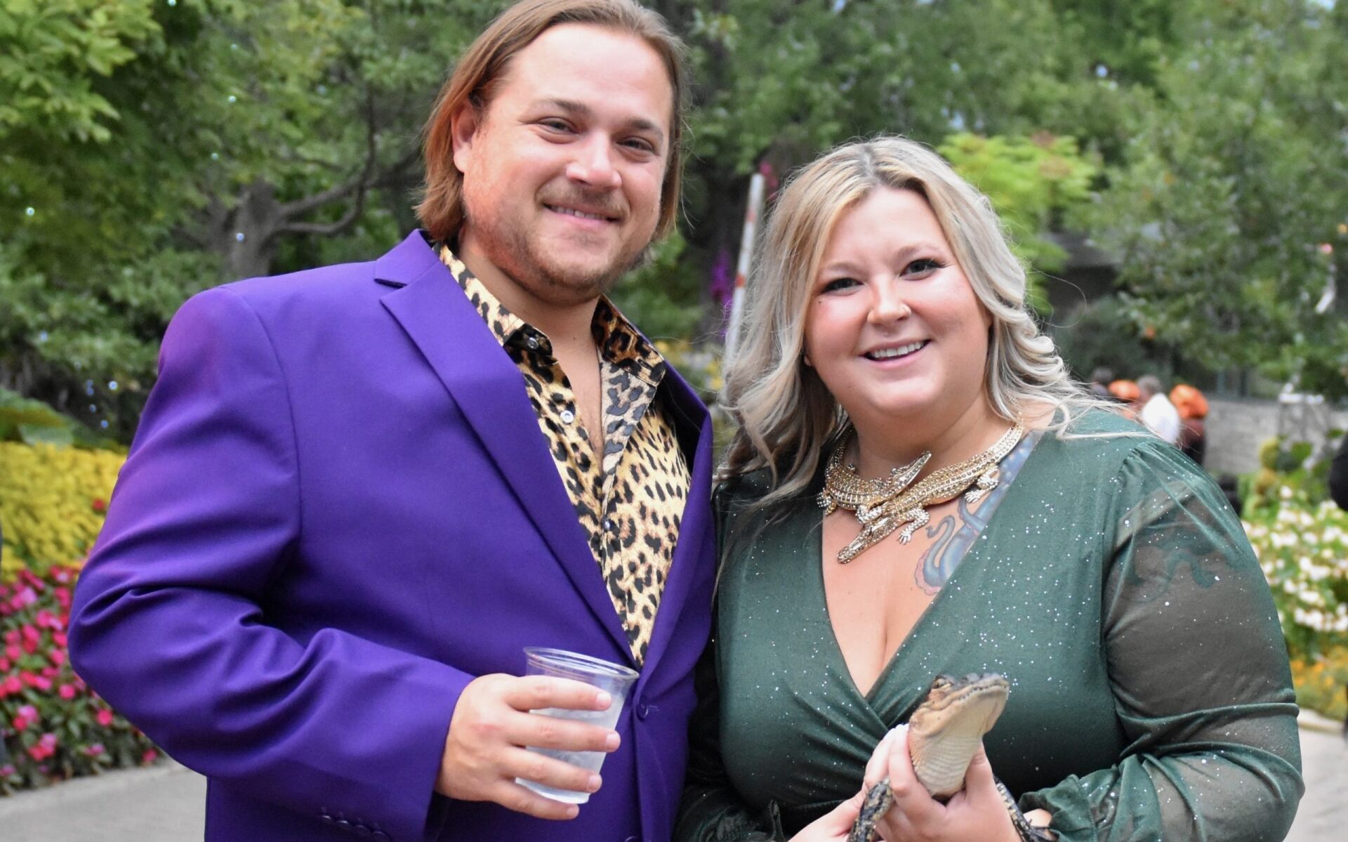 man on the left wearing blue jacket next to women on teh rghin green dress, and holding a small abby alligator durng an outdoor event. Green bushes in background.